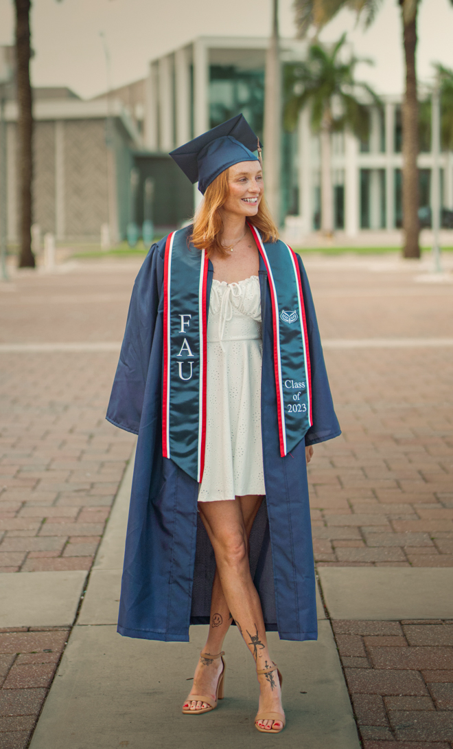 Female college graduate walking on campus wearing a navy cap and gown with a custom embroidered graduation stole, demonstrating high-quality satin grad stoles and custom graduation stoles from The Sash Company.